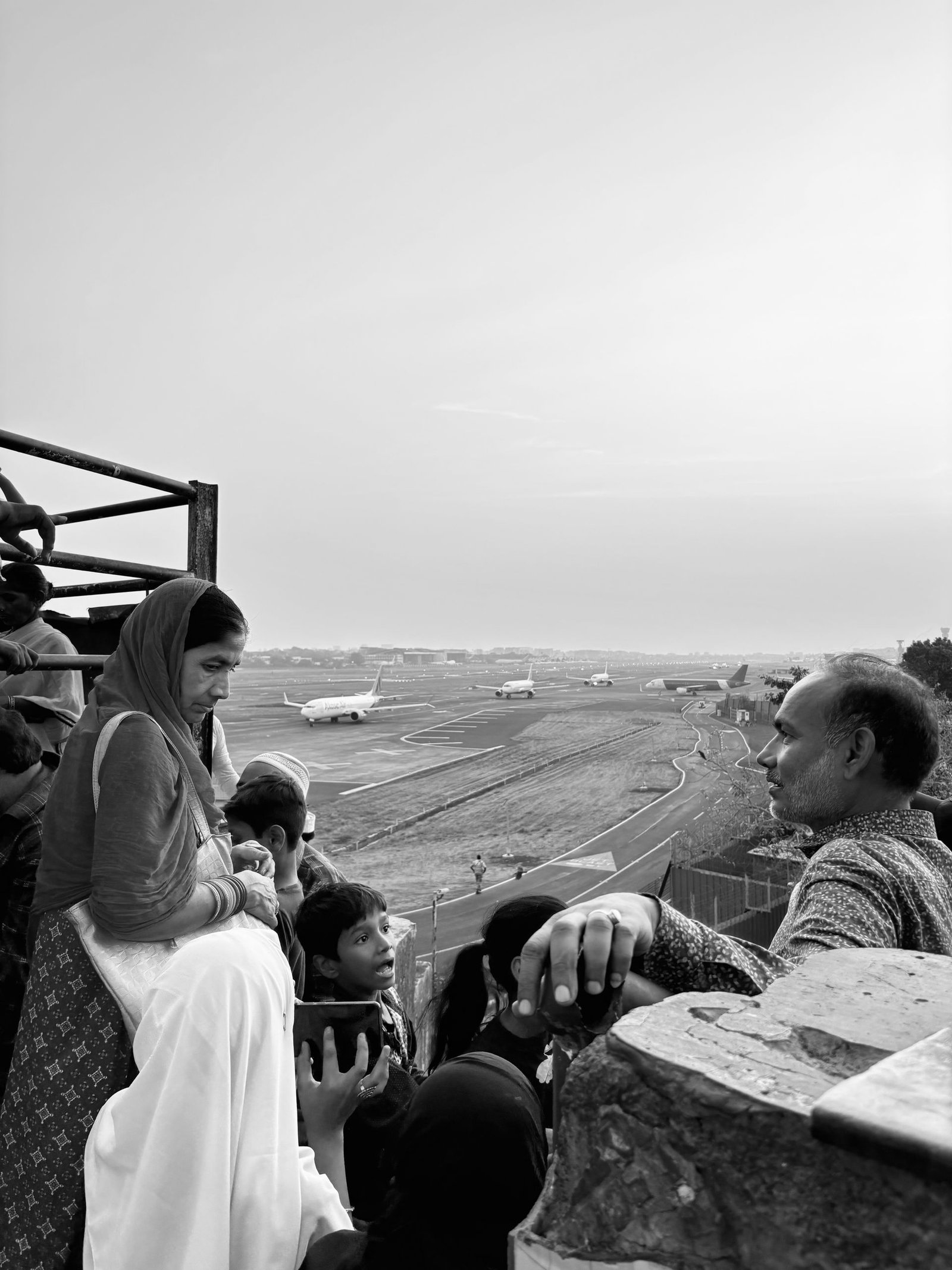 Families gather at a Mumbai airport viewing gallery, capturing a candid moment with planes in the background.